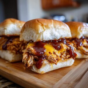 Three BBQ chicken sliders topped with melted cheese and BBQ sauce on a wooden board.