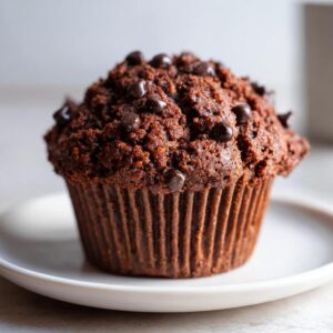 A single chocolate zucchini muffin on a white plate, topped with chocolate chips.