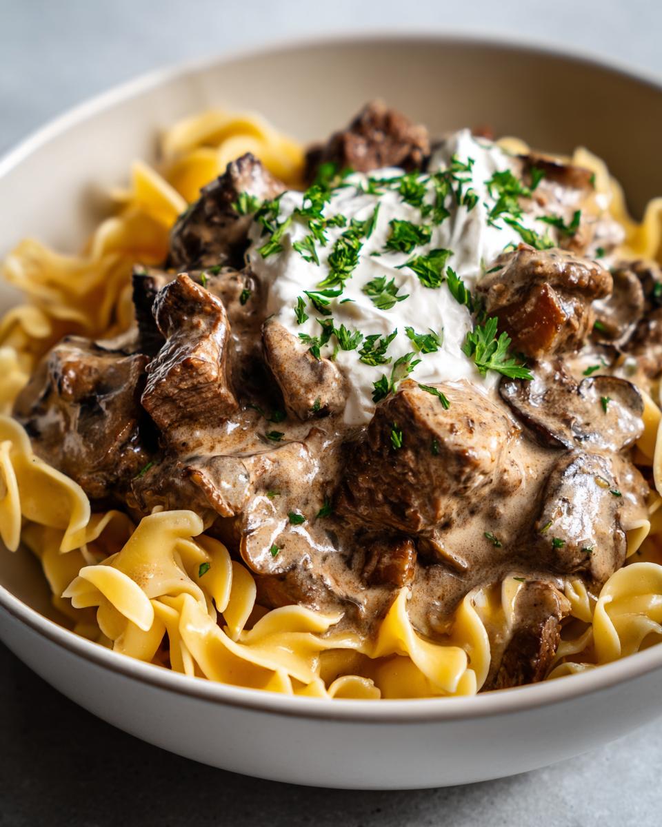 A bowl of creamy Beef Stroganoff with egg noodles, topped with sour cream and fresh parsley.