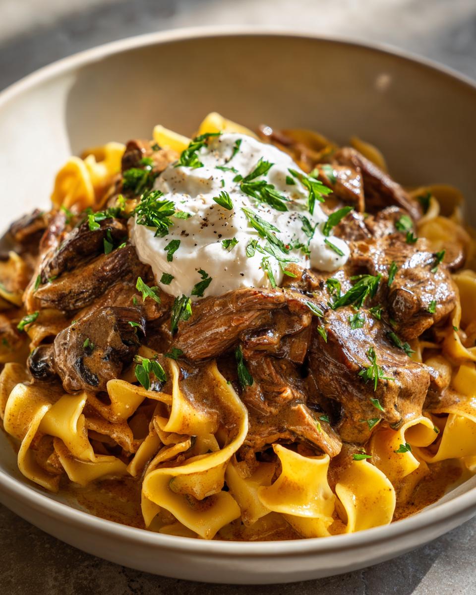 A bowl of creamy Beef Stroganoff with egg noodles, topped with sour cream and fresh parsley.