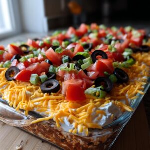 A close-up view of a freshly made 7 Layer Dip in a glass baking dish, topped with shredded cheese, tomatoes, olives, and green onions.