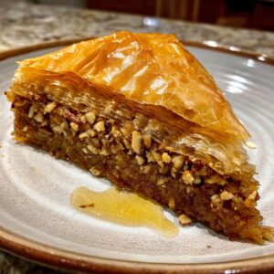 A close-up of a golden-brown slice of Baklava on a plate, showcasing layers of flaky pastry and chopped nuts, with a drizzle of syrup.