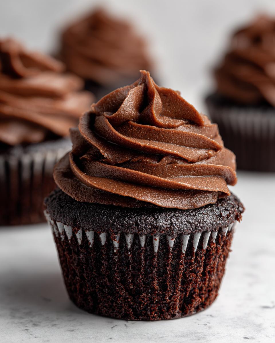 Close-up of a decadent chocolate cupcake topped with creamy chocolate frosting, with other chocolate cupcakes blurred in the background.