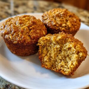 Three golden brown oatmeal muffins on a white plate, with one muffin broken in half to show its fluffy interior.