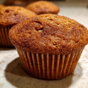 Close-up of a fluffy pumpkin muffin in a brown paper liner, with other muffins blurred in the background.