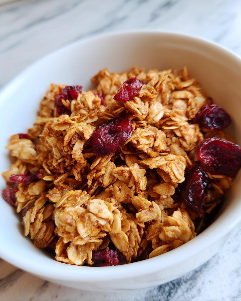 A close-up of a white bowl filled with homemade granola and dried cranberries, showcasing crunchy clusters.