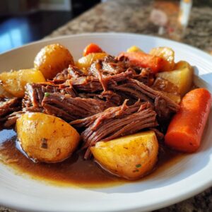 A close-up of tender, shredded slow cooker pot roast served with golden potatoes and bright orange carrots in a rich brown gravy.