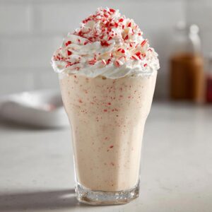 A tall glass filled with a creamy Candy Cane Milkshake, topped high with whipped cream and crushed peppermint pieces.