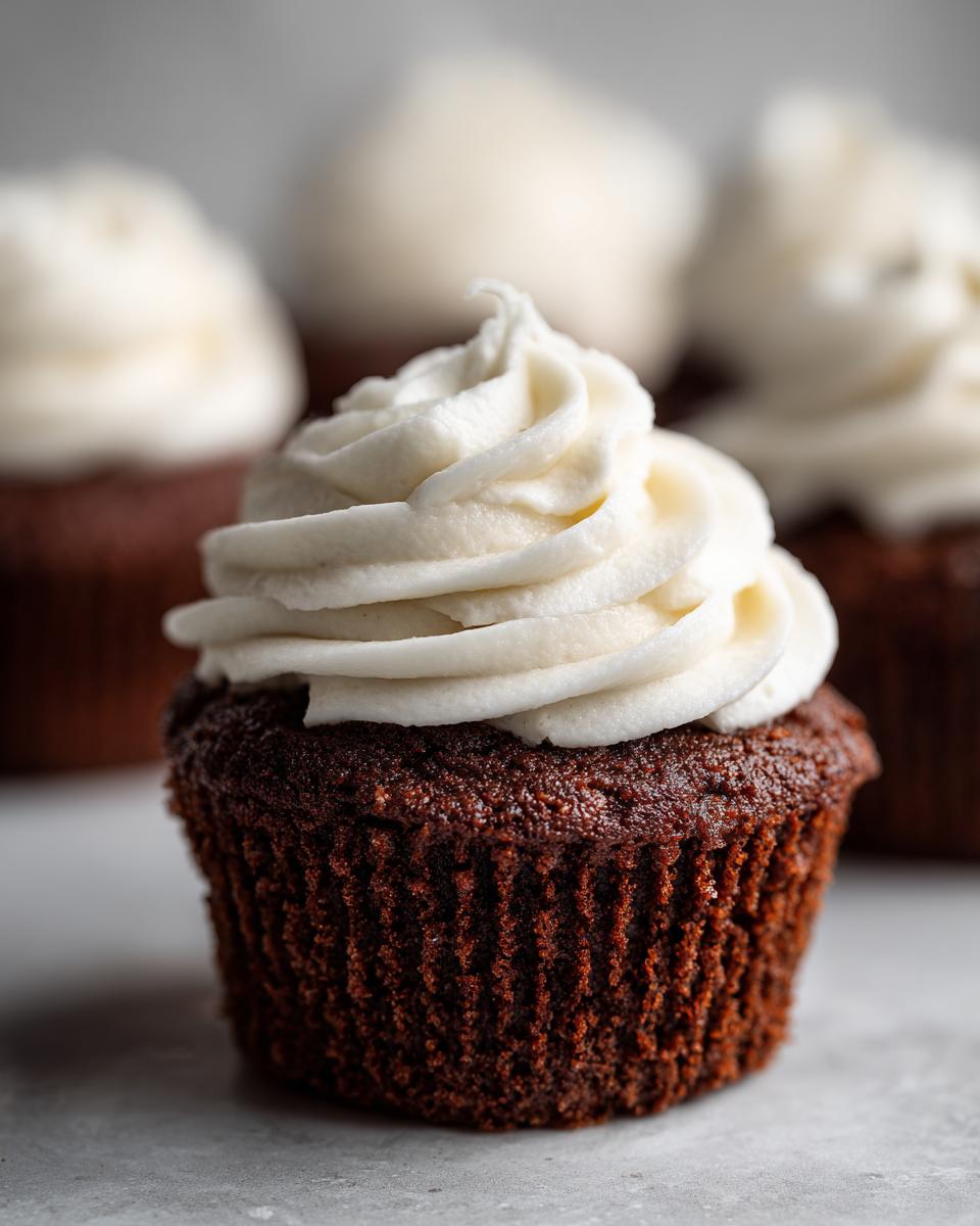 A single, rich, dark brown gingerbread cupcake topped with a swirl of white cream cheese frosting, with more cupcakes blurred in the background.