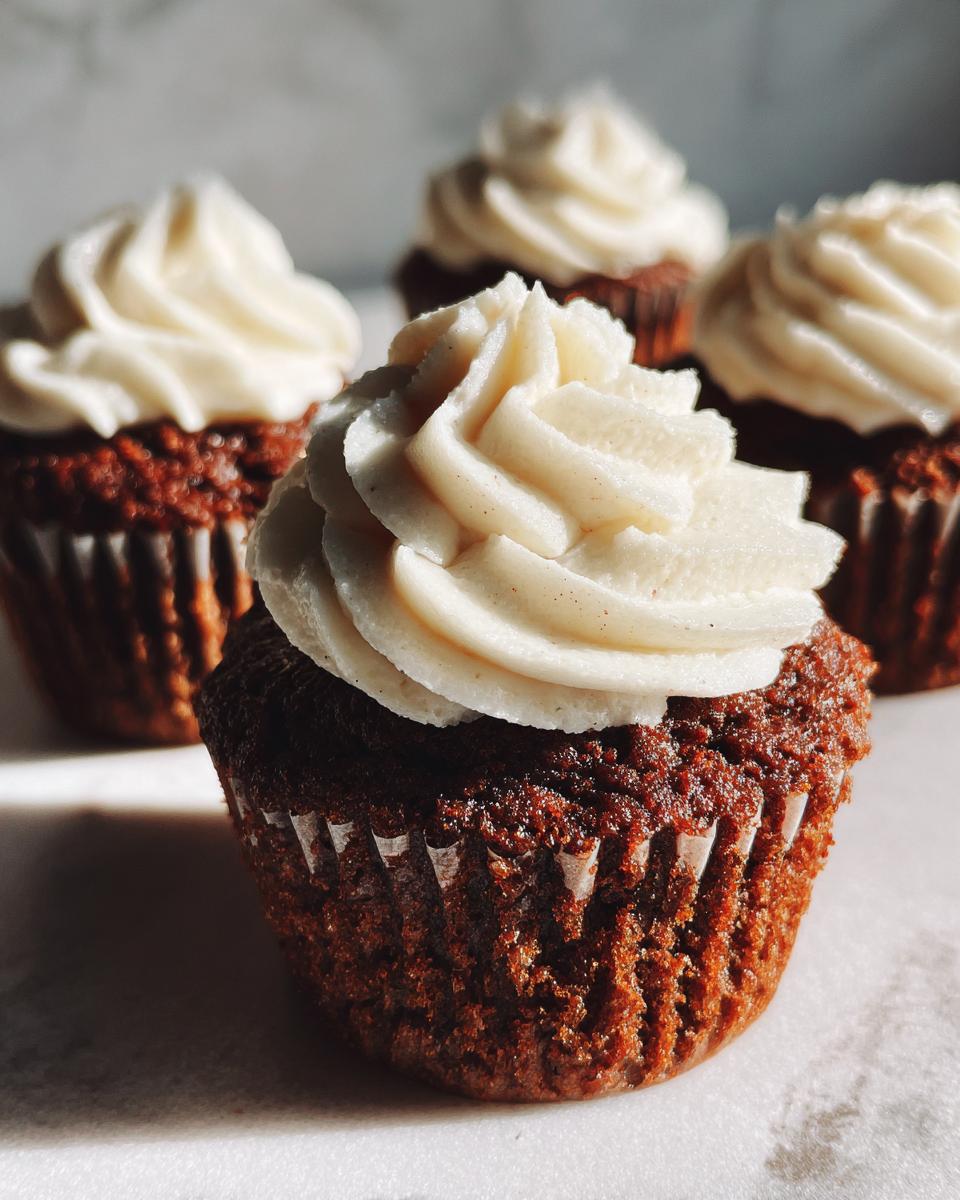 A close-up, macro shot of one perfectly frosted Gingerbread Cupcakes in the foreground with three others blurred behind it.
