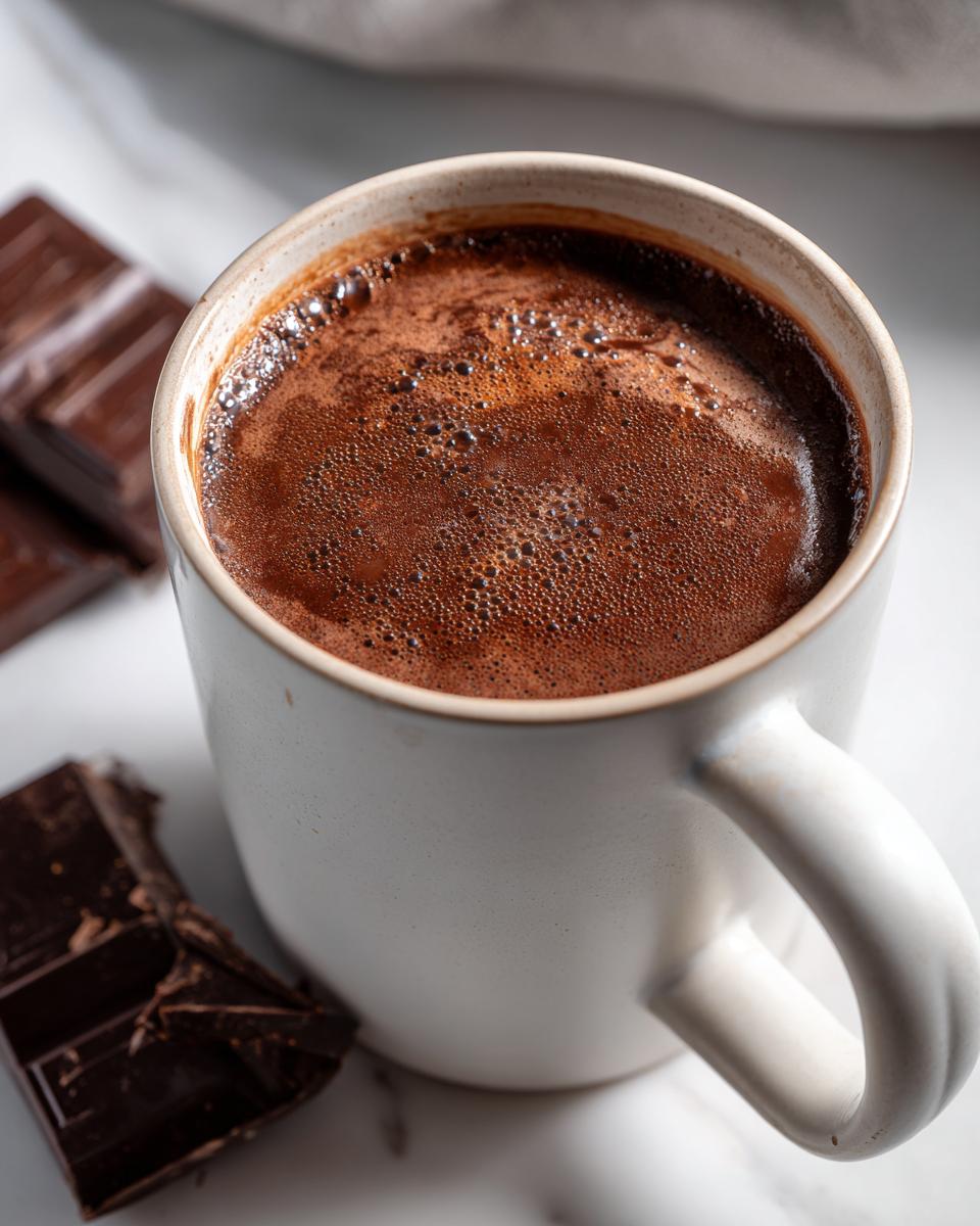 Close-up of a mug filled with thick, dark brown Champurrado, showing a frothy top, next to pieces of dark chocolate.