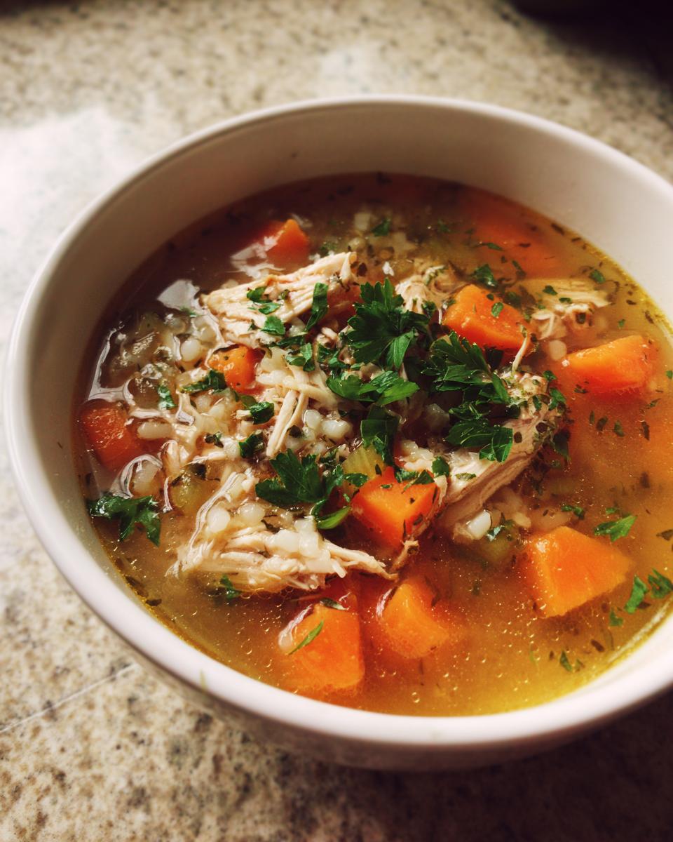 A close-up of a bowl of comforting turkey soup with shredded turkey, diced carrots, barley, and fresh parsley.