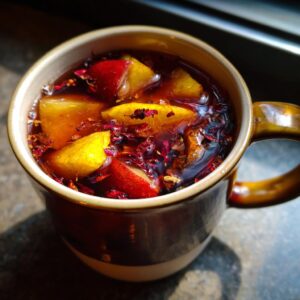 Close-up of a warm mug filled with rich Ponche Navideño, featuring chunks of apple and orange floating with dried red spices.