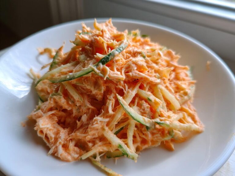 A close-up of a creamy, orange-hued kani salad mixed with shredded cucumber strips on a white plate.