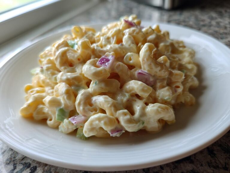 A close-up serving of creamy Amish macaroni salad featuring elbow pasta, red onion, and celery on a white plate.